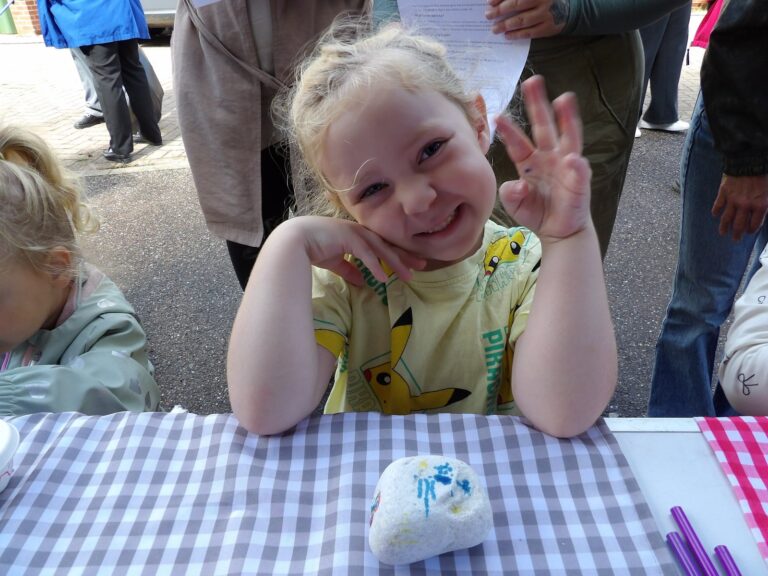 A young girl waving at a craft table with a decorated stone in front of her