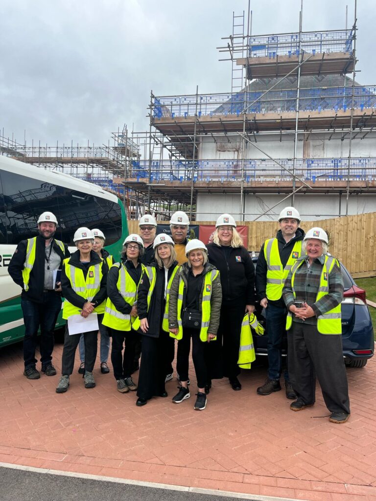 A large group in hard hats and hi-vis vests standing in front of homes under construction and covered in scaffolding