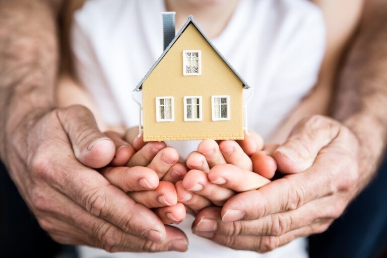 A pair of hands cupped around a small wooden model house, held by another pair of hands