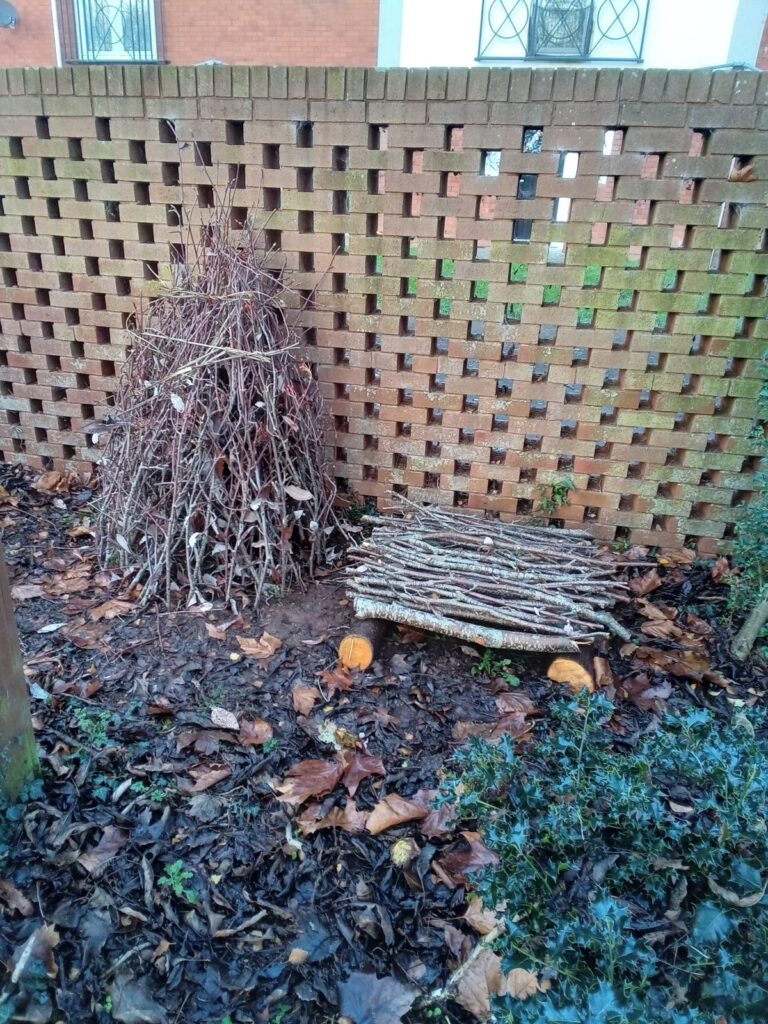 An insect refuge built from sticks and cuttings standing against a brick wall in a communal garden