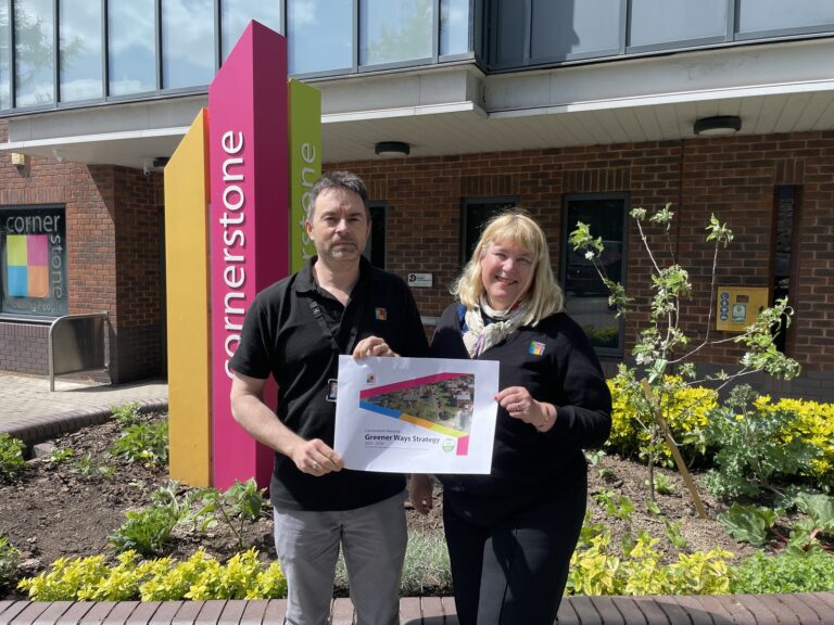 Two Cornerstone colleagues holding a printed copy of the Greener Ways Strategy beside the office sign