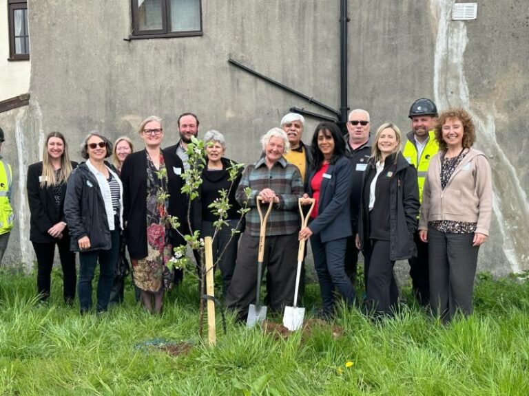 A group of Cornerstone staff and residents standing around a newly planted tree, two of them holding spades
