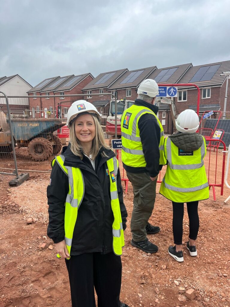 A Cornerstone staff member in a hi-vis jacket on a construction site, with colleagues and newly built homes behind her