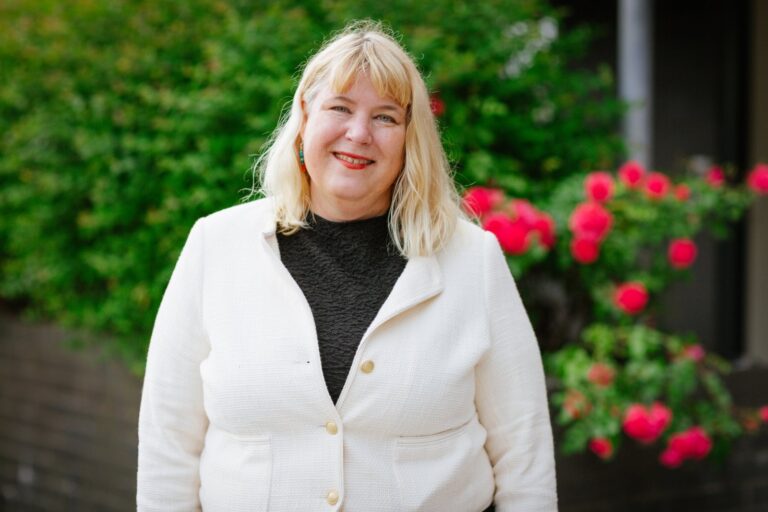 A Cornerstone colleague in a cream jacket smiling, standing outdoors beside a climbing rose