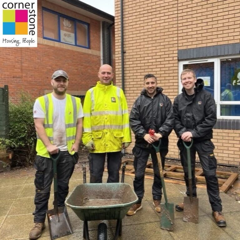 Four colleagues with spades and a wheelbarrow outside a community building